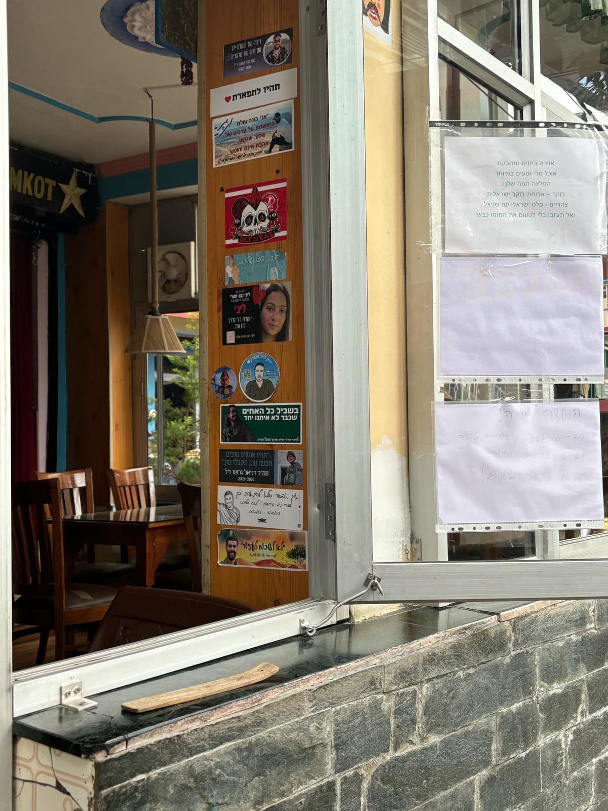 An open window is shown, and inside the window, a wall is covered in Hebrew stickers with images of soldiers.